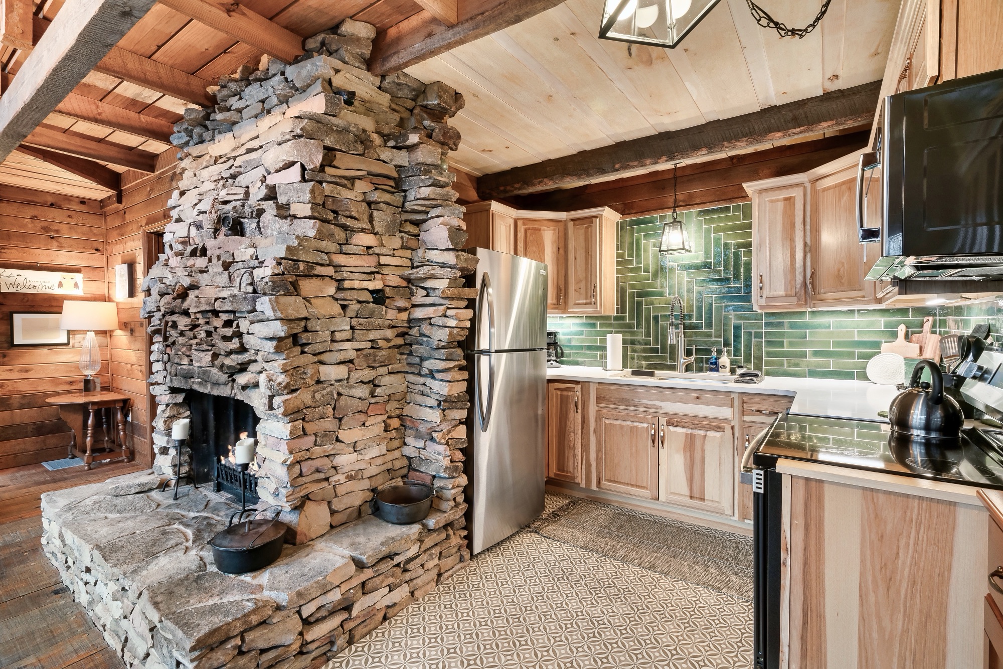 Stone chimney and fireplace beside the kitchen's green tile backsplash and stainless refrigerator